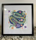 Framed folk art print featuring a green snake coiled on a dark blue circular background, surrounded by colorful flowers, displayed in a black frame on a granite countertop.