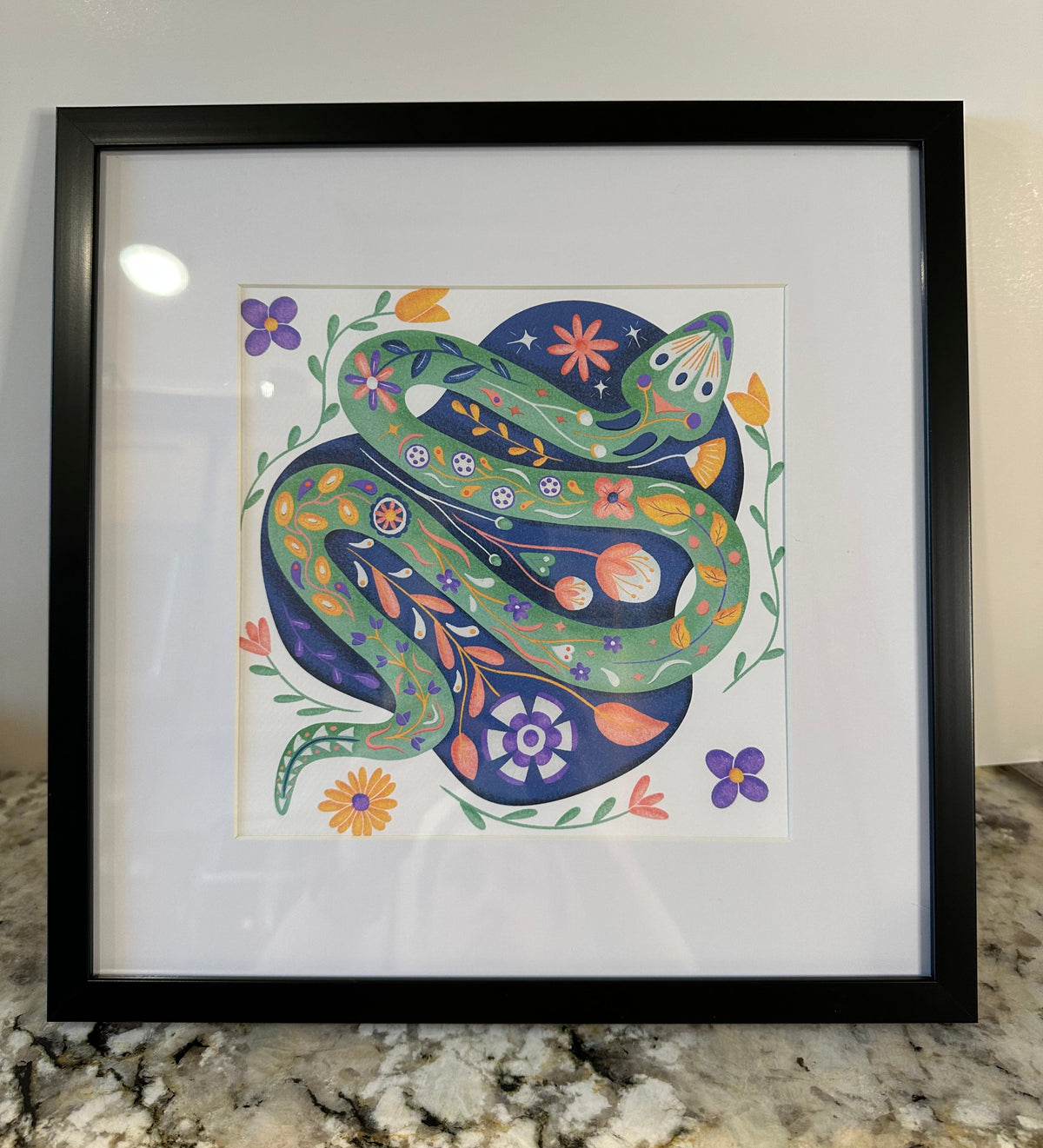Framed folk art print featuring a green snake coiled on a dark blue circular background, surrounded by colorful flowers, displayed in a black frame on a granite countertop.